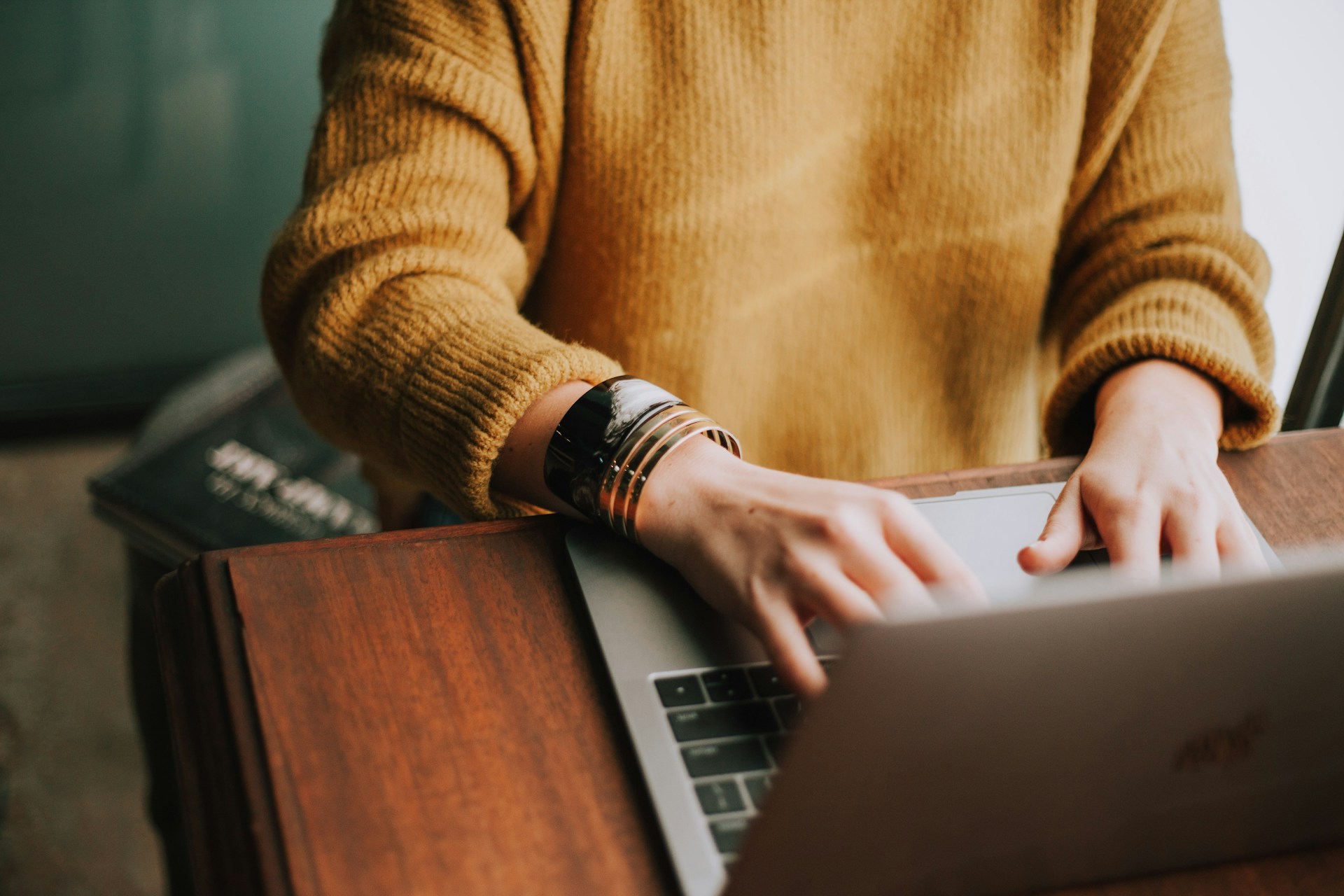 A person in a knitted jumper typing on a laptop at a wooden desk.